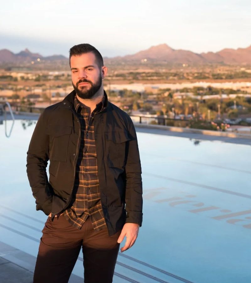 A man in a jacket and plaid shirt stands by a pool with a scenic view of mountains and a city in the background at sunset.